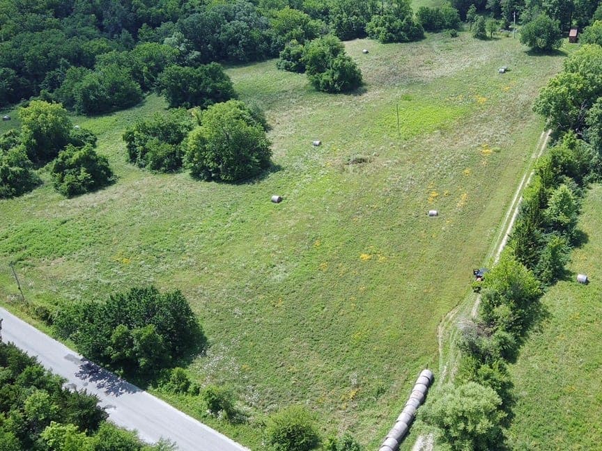 Aerial drone photo of a sloped green hayfield with round hay bales scattered across it, bordered by a road on one side and a tree line on the other